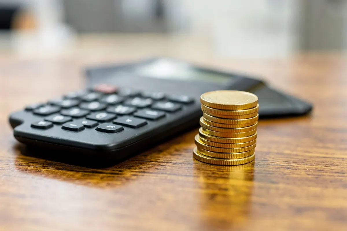 Uma calculadora e uma pilha de moedas de ouro em uma mesa de madeira, plano detalhe, luz de escritório vinda de cima, sem ação.