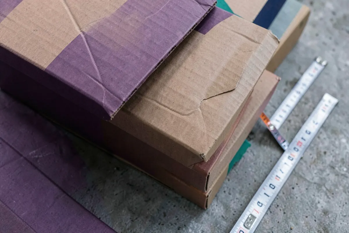 Pilha de caixas de papelão de tamanhos variados em um chão de cimento, com uma fita métrica ao lado.