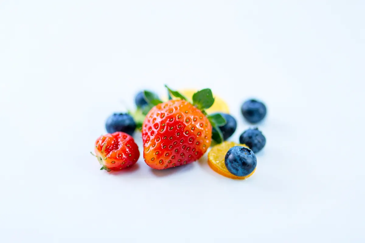 Um close-up de frutas frescas como morangos, mirtilos e fatias de laranja dispostas artisticamente em uma superfície branca.