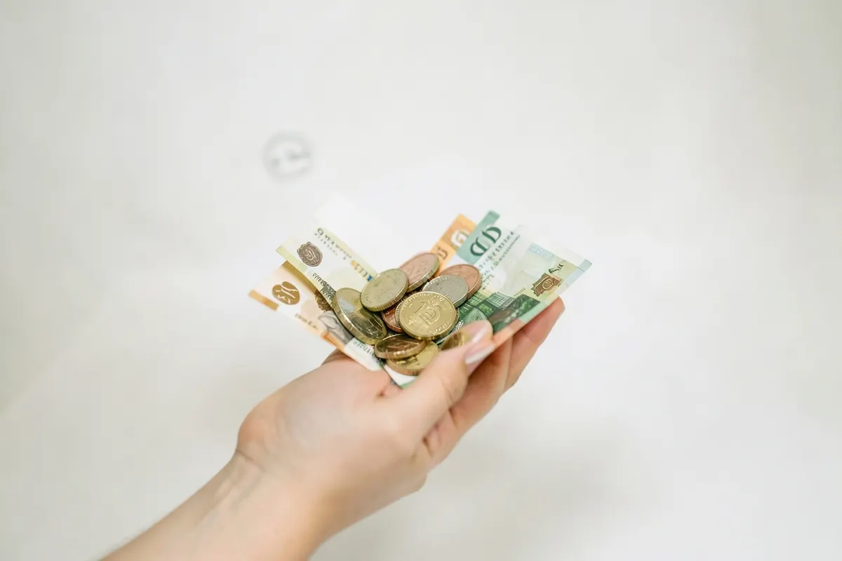 Uma mão segurando moedas e notas de dinheiro, com um ícone de pagamento ao fundo.