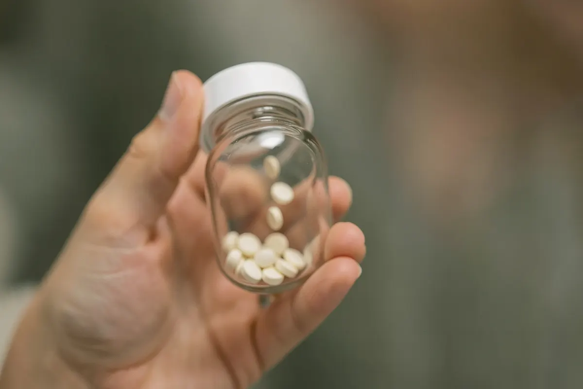 Close-up de uma mão segurando um frasco de comprimidos com algumas pílulas caindo dele.