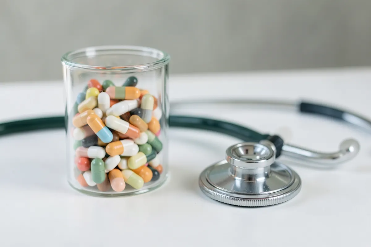 Uma caixa de medicamentos com vários comprimidos e cápsulas, ao lado de um estetoscópio.