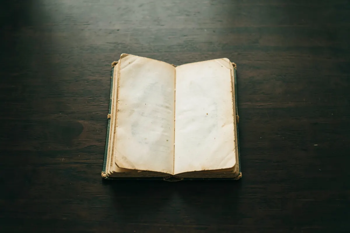 Um close-up de um livro antigo com a capa desgastada, com páginas amareladas visíveis, sobre uma mesa de madeira escura. A luz suave vem de cima, iluminando o centro do livro.