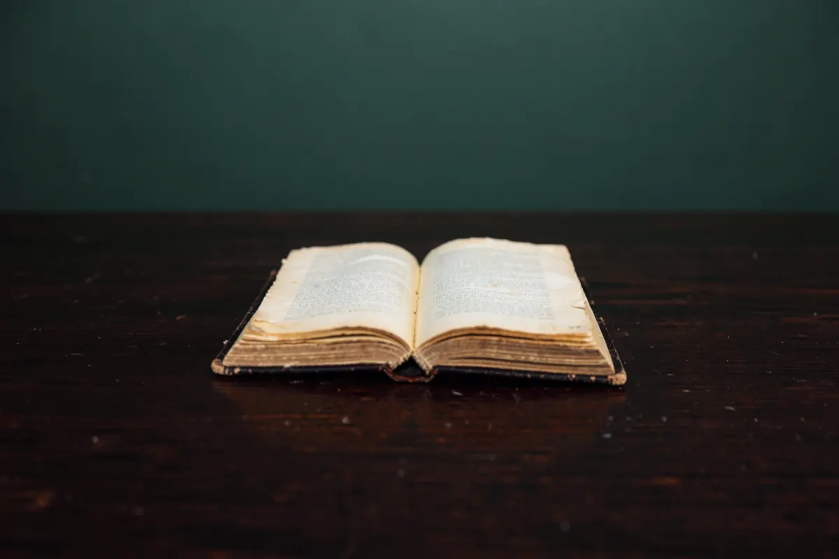 Um close-up de um livro antigo com a capa desgastada, com páginas amareladas visíveis, sobre uma mesa de madeira escura. A luz suave vem de cima, iluminando o centro do livro.
