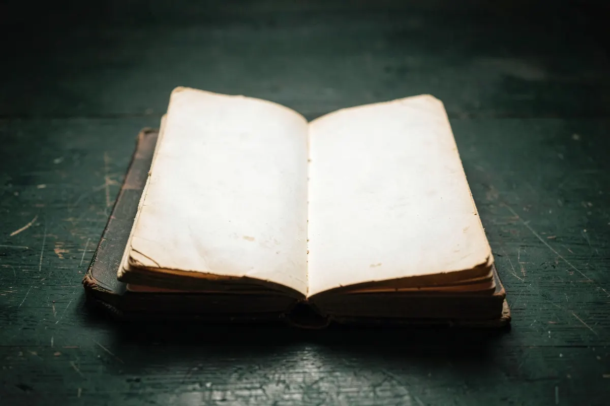 Um close-up de um livro antigo com a capa desgastada, com páginas amareladas visíveis, sobre uma mesa de madeira escura. A luz suave vem de cima, iluminando o centro do livro.