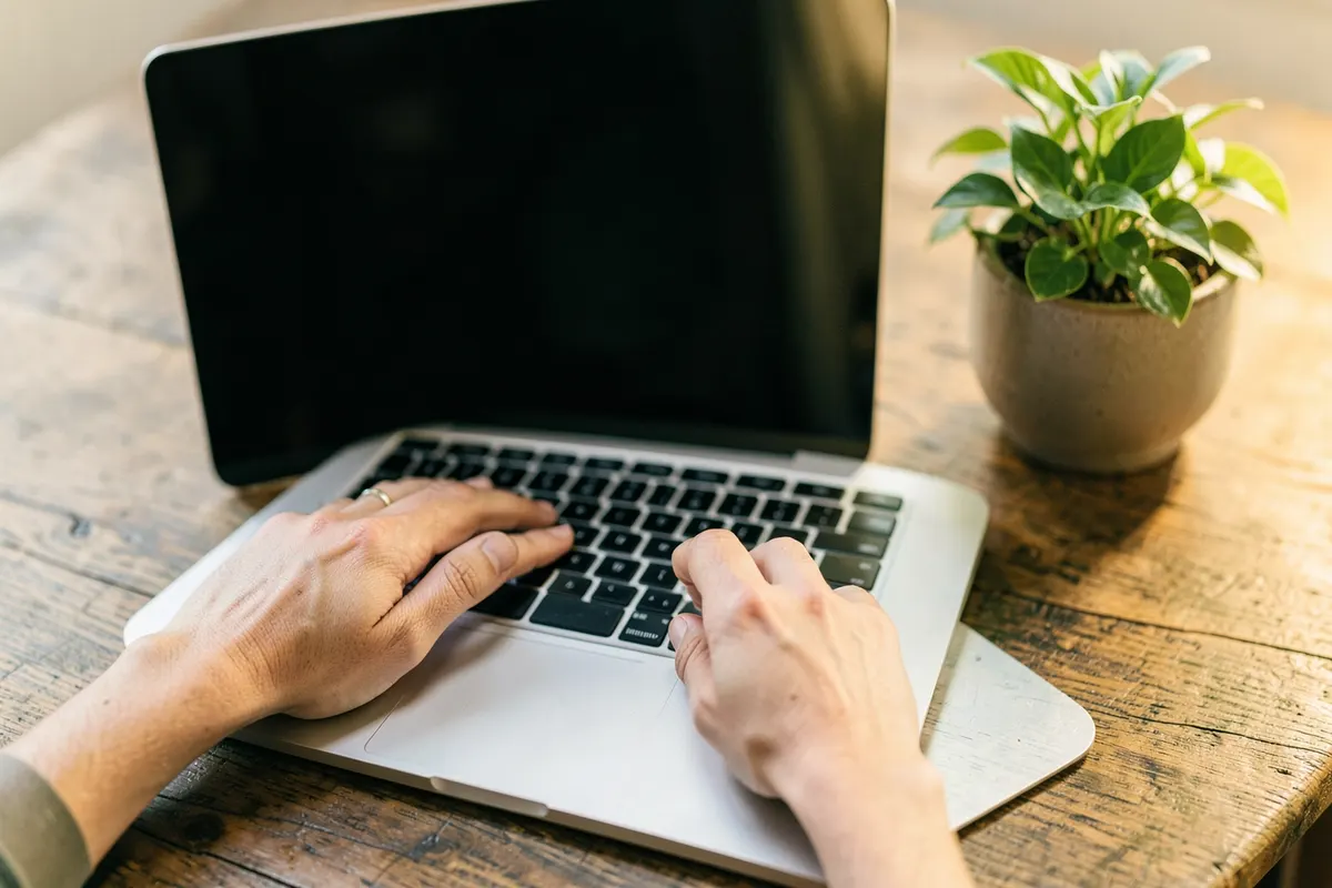Mão digitando em um laptop com uma tela em branco, em uma mesa de madeira com um pequeno vaso de planta.